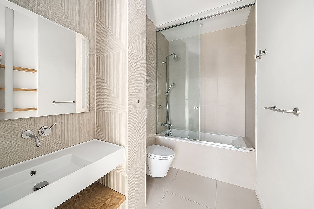 This modern, minimalist bathroom features a sleek wall-mounted vanity with a white basin and a large, integrated mirror. The space is finished with textured, light-toned wall tiles and a glass-enclosed shower-tub combination, creating a clean and airy atmosphere. The wall-hung toilet and neutral color palette contribute to a contemporary, high-end aesthetic.