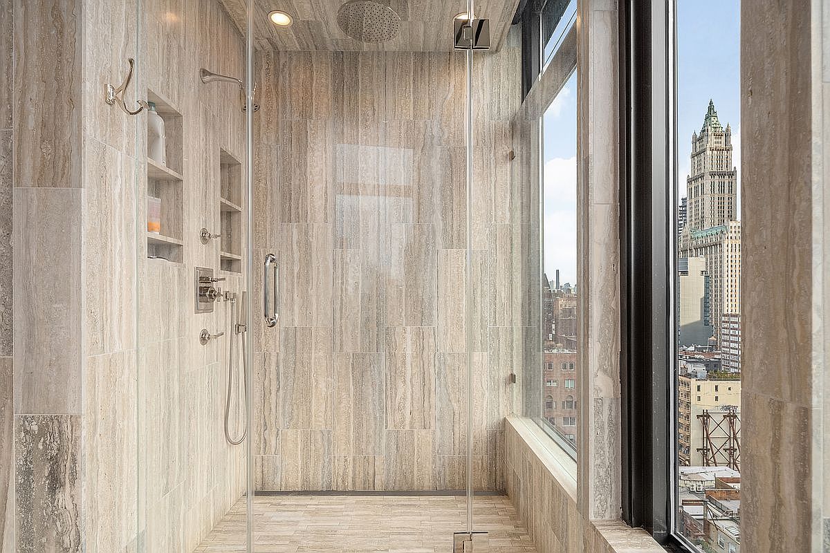 This luxurious, modern walk-in shower features floor-to-ceiling beige travertine tile and a seamless glass enclosure. The space is highlighted by a stunning, unobstructed view of the iconic Woolworth Building through a large floor-to-ceiling window. The design includes high-end fixtures, a built-in recessed shelf, and a rainfall showerhead, creating a sophisticated and serene spa-like atmosphere.