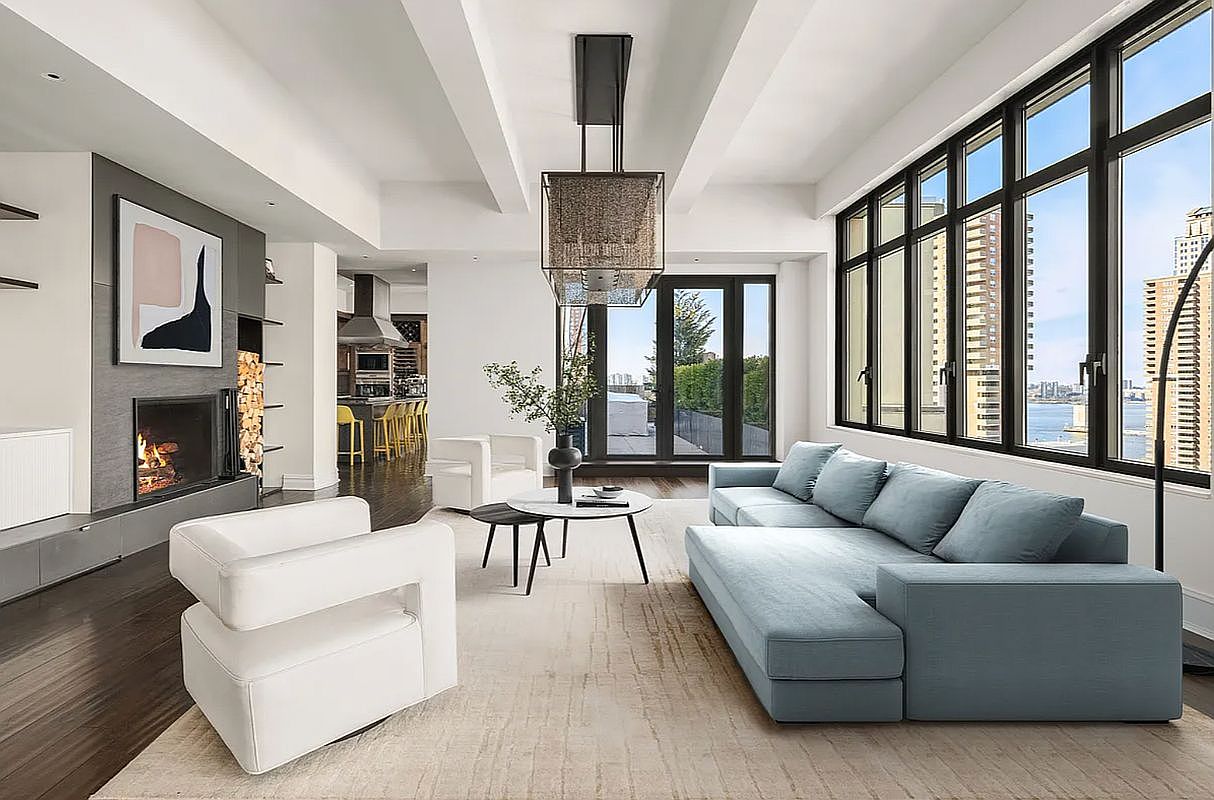 This sophisticated, open-concept living room features high ceilings with architectural beams, a sleek fireplace, and a large, light-blue sectional sofa. Expansive floor-to-ceiling windows flood the space with natural light while offering panoramic city and water views. The room is anchored by a neutral area rug and modern white armchairs, creating a luxurious and airy atmosphere perfect for high-end urban living.