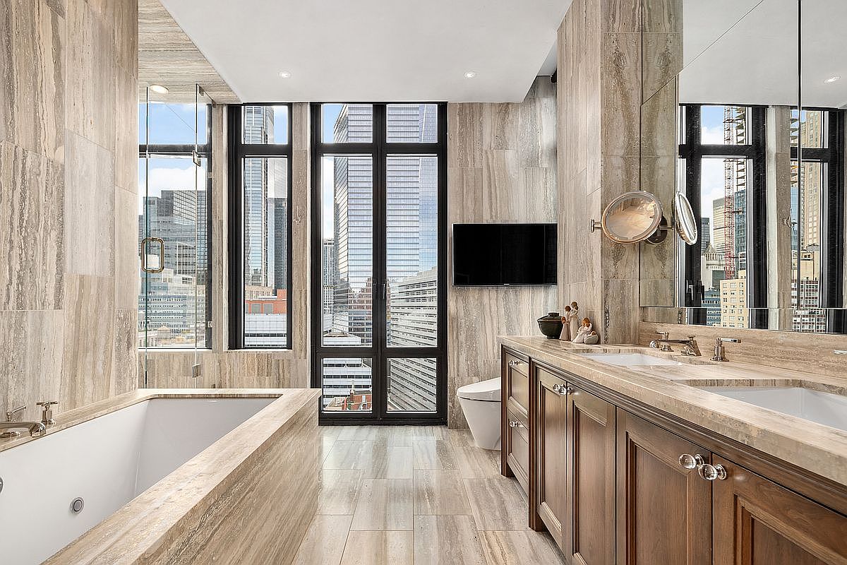 This luxurious primary bathroom features floor-to-ceiling travertine stone tiling and a deep soaking tub positioned next to expansive windows that offer a dramatic urban skyline view. The space includes a double vanity with rich wood cabinetry, dual sinks, and a wall-mounted television, creating a sophisticated and serene spa-like atmosphere. The perspective is centered, highlighting the seamless integration of high-end materials and the impressive city backdrop.