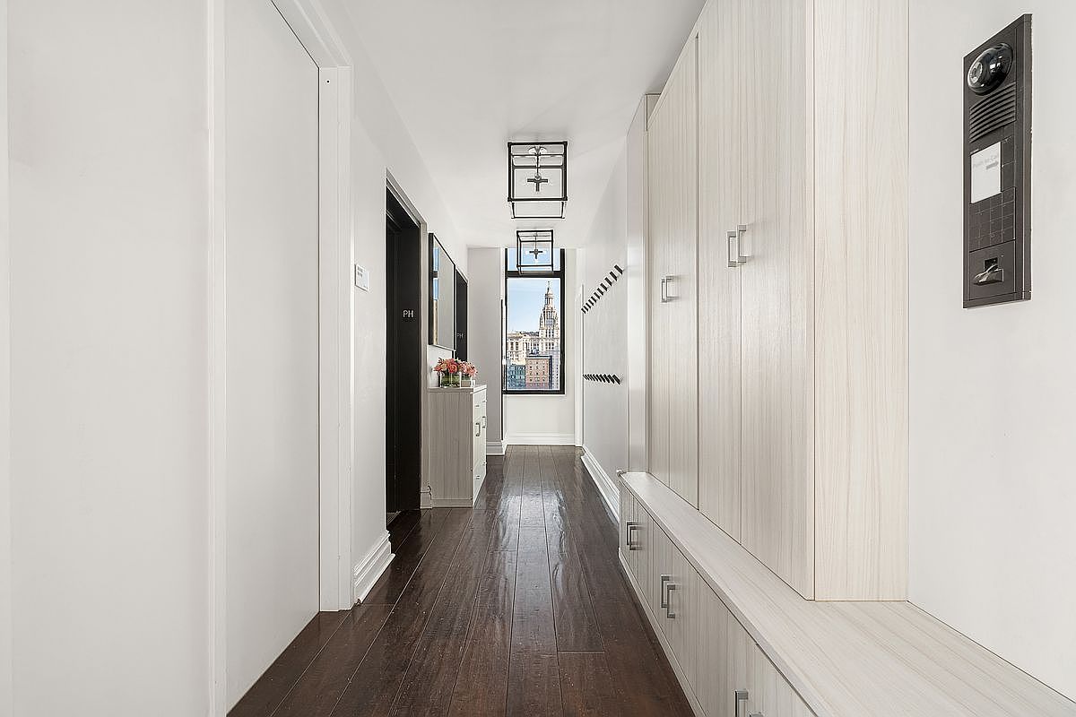 This bright, modern hallway features dark hardwood flooring that contrasts beautifully with the crisp white walls and custom light-wood cabinetry. The space is illuminated by two stylish, geometric pendant lights and leads to a window at the end, which provides a clear view of the city skyline. The overall aesthetic is clean, sophisticated, and highly functional, offering ample storage solutions.