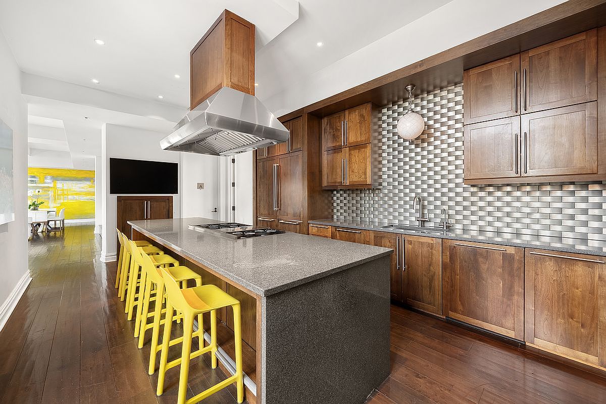 This modern kitchen features rich, dark wood cabinetry paired with a sleek, speckled grey stone island and countertops. A striking metallic tile backsplash adds texture, while bright yellow bar stools provide a vibrant pop of color against the warm hardwood floors. The space is illuminated by recessed lighting and a stylish pendant light, creating a sophisticated and inviting atmosphere.
