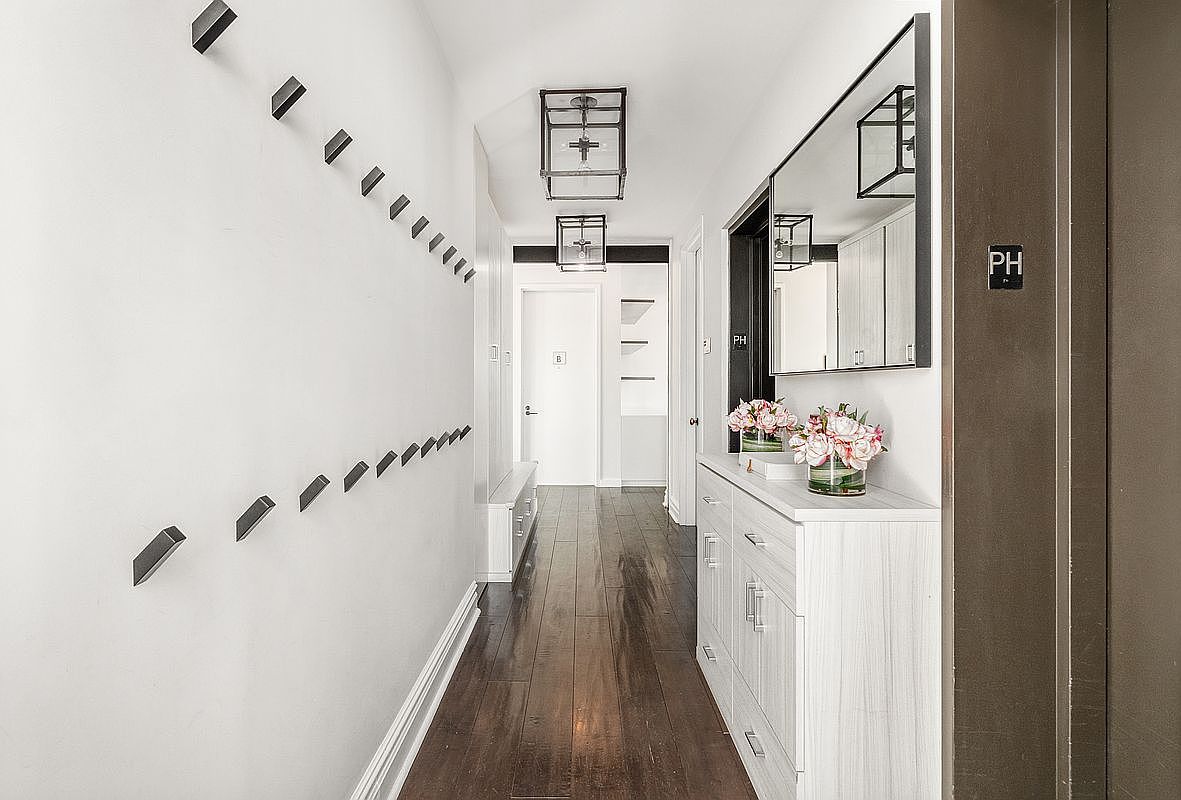 This elegant, modern hallway features dark hardwood flooring that contrasts beautifully with crisp white walls and cabinetry. The space is accented by a series of minimalist wall-mounted hooks, two geometric pendant light fixtures, and a white console table topped with a floral arrangement and a large mirror. The perspective is a straight-on, eye-level shot looking down the corridor toward a white door, creating a clean, sophisticated, and welcoming atmosphere.