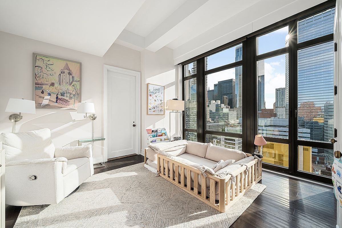 This bright and airy nursery or child's bedroom features a modern aesthetic with floor-to-ceiling windows offering expansive city skyline views. The room is furnished with a comfortable white armchair, a wooden toddler bed with a soft mattress, and a textured area rug, creating a cozy and inviting atmosphere. The neutral color palette and clean lines emphasize the high-end, contemporary design of the space.