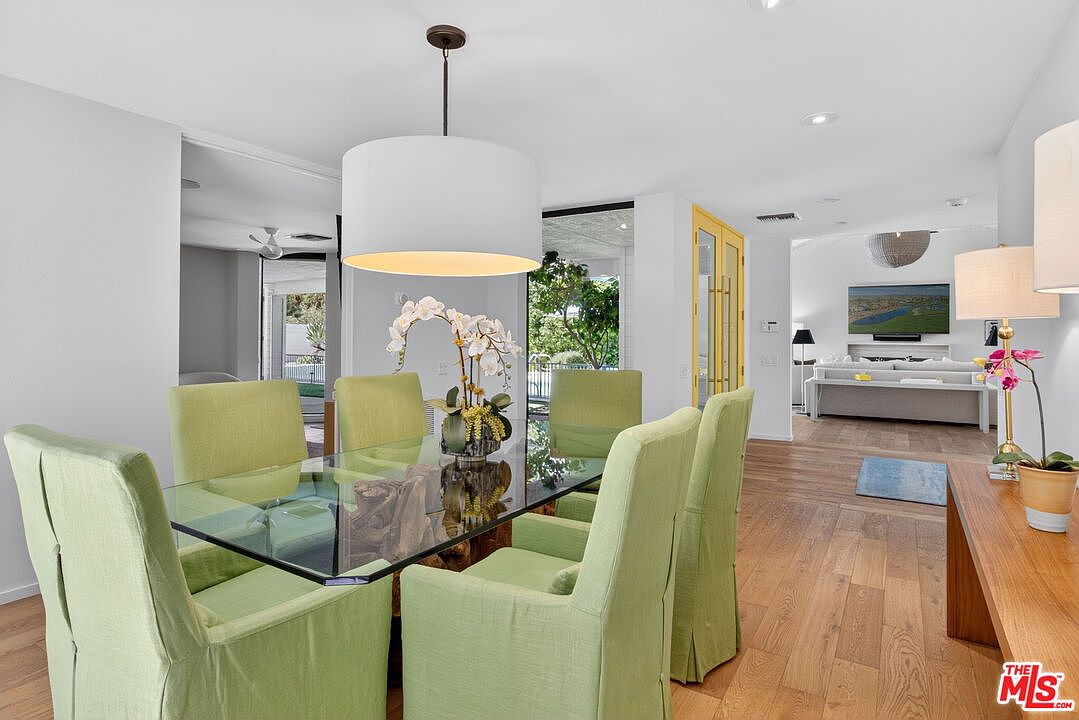 This bright dining area features a glass-topped table surrounded by six light green upholstered chairs, centered beneath a large white drum pendant light. The room flows seamlessly into an adjacent living space, which is visible through an open doorway and features a television mounted above a fireplace. The interior is characterized by light wood flooring, neutral walls, and a clean, contemporary aesthetic that emphasizes an open-concept layout.