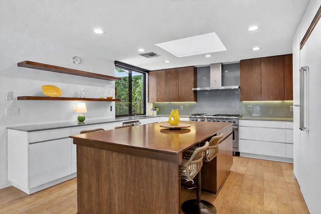 This modern kitchen features a large central island with a wood-toned base and a spacious countertop, complemented by two woven bar stools. The space is defined by a mix of white lower cabinetry and warm wood-toned upper cabinets, accented by open shelving and a sleek stainless steel range hood. A large window above the sink area brings in natural light, while recessed lighting and a skylight brighten the entire room.