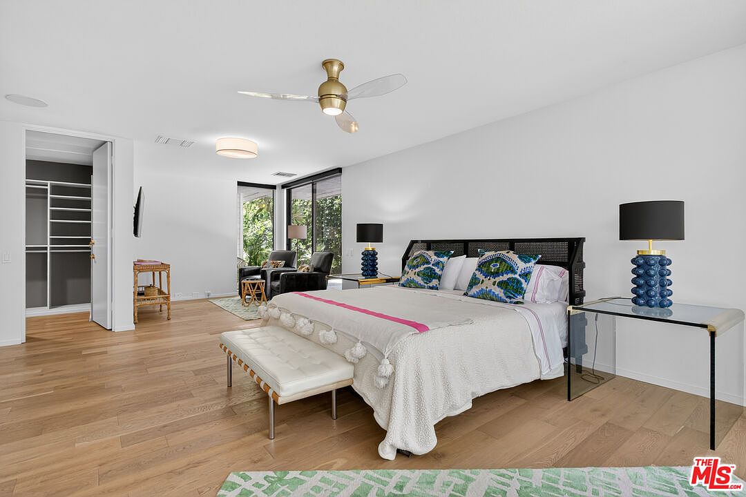 This spacious primary bedroom features light wood flooring and a neutral color palette, anchored by a large bed with a dark, textured headboard and vibrant patterned accent pillows. A white bench sits at the foot of the bed, while a glass-topped side table with a blue textured lamp adds a touch of modern elegance. Large sliding glass doors provide natural light and a view of the lush greenery outside, creating a serene and inviting atmosphere.