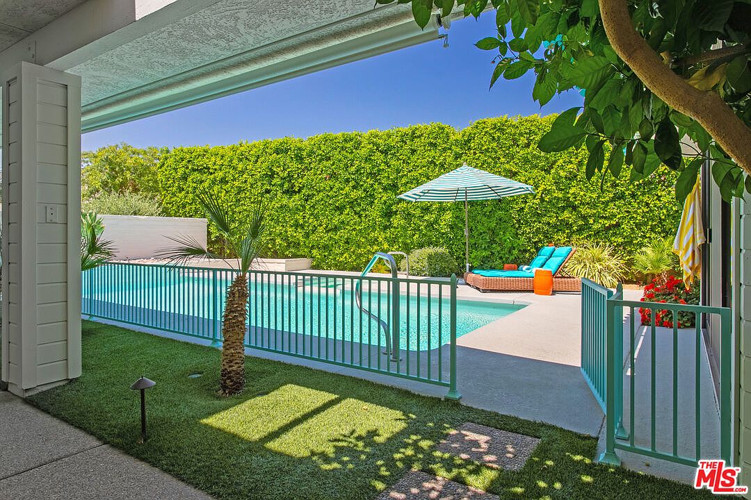 This bright, sun-drenched backyard features a sparkling swimming pool enclosed by a teal-colored metal safety fence. A comfortable lounge area with a striped umbrella and wicker furniture sits on the concrete patio, while a lush, tall green hedge provides complete privacy for the space. The perspective is captured from a covered patio area, looking out toward the pool and the vibrant landscaping.