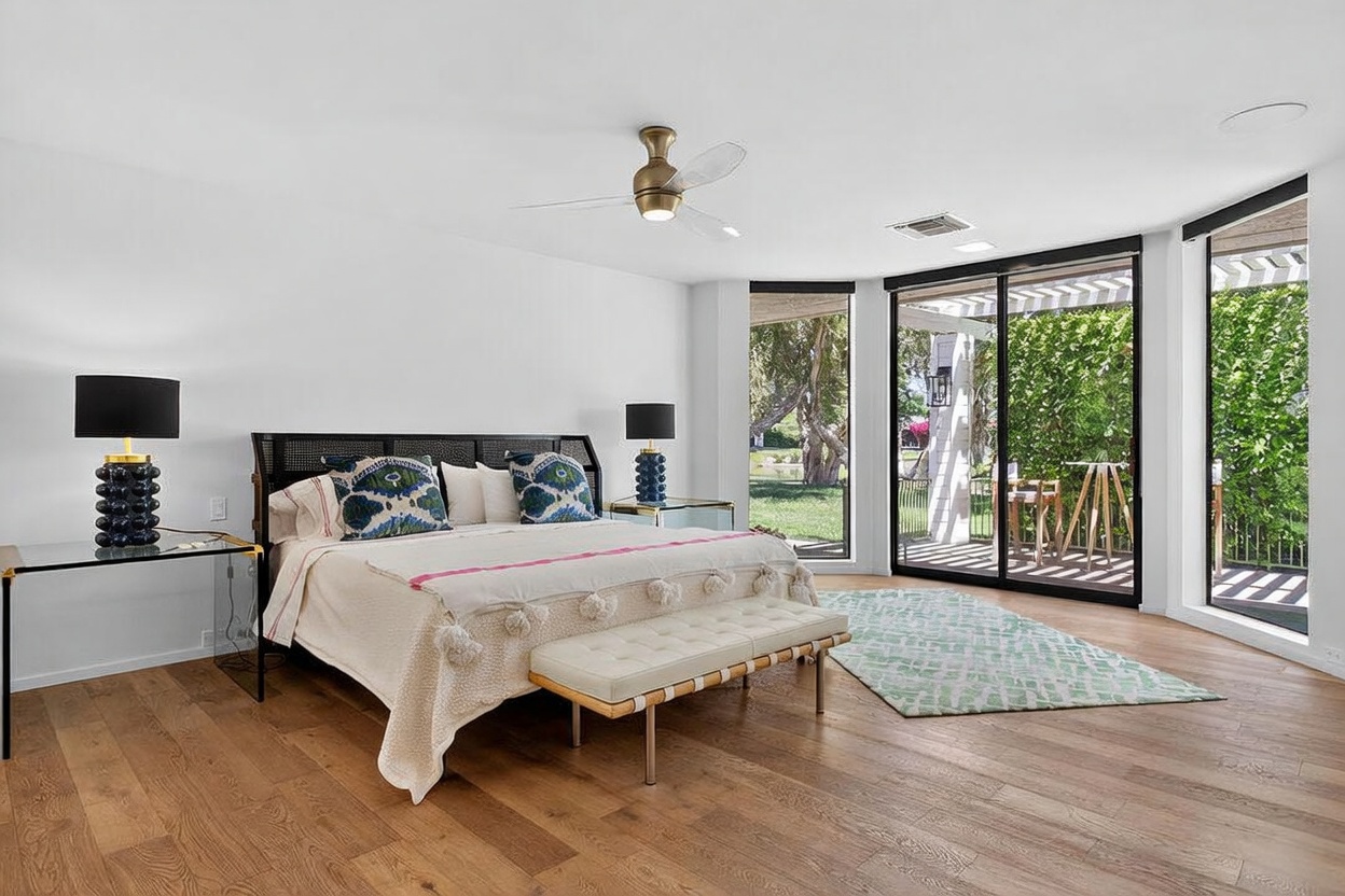 This spacious primary bedroom features light wood flooring, white walls, and a large bed with a dark, textured headboard. Large glass sliding doors provide a seamless transition to an outdoor patio, allowing for ample natural light and views of the surrounding greenery. A modern ceiling fan and matching bedside lamps with unique, stacked-sphere bases complete the contemporary aesthetic.