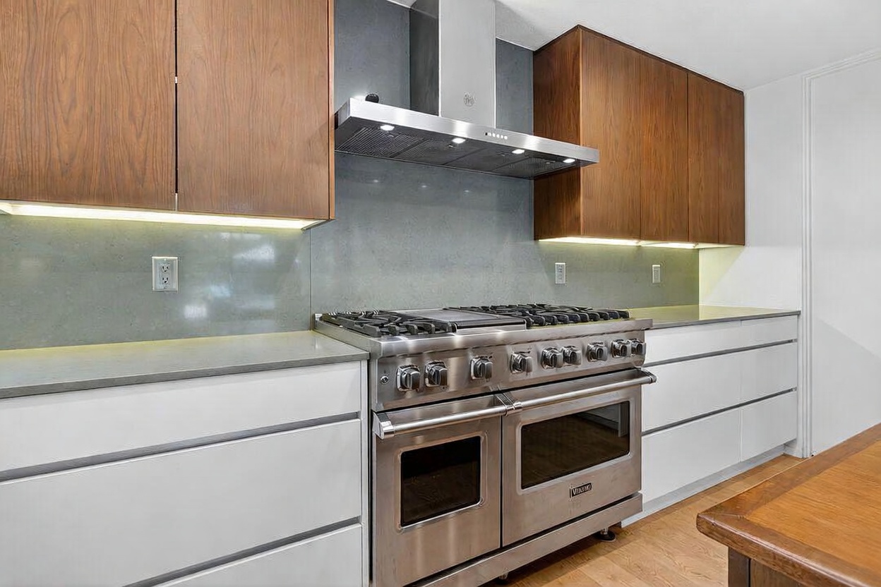 This modern kitchen features a professional-grade stainless steel range with a matching range hood centered against a sleek, solid-surface backsplash. The space is defined by a clean, minimalist aesthetic with warm-toned wood upper cabinets contrasting against crisp white lower cabinetry and light-colored countertops. The perspective is a straight-on eye-level shot, highlighting the high-end appliances and the seamless integration of the cabinetry.
