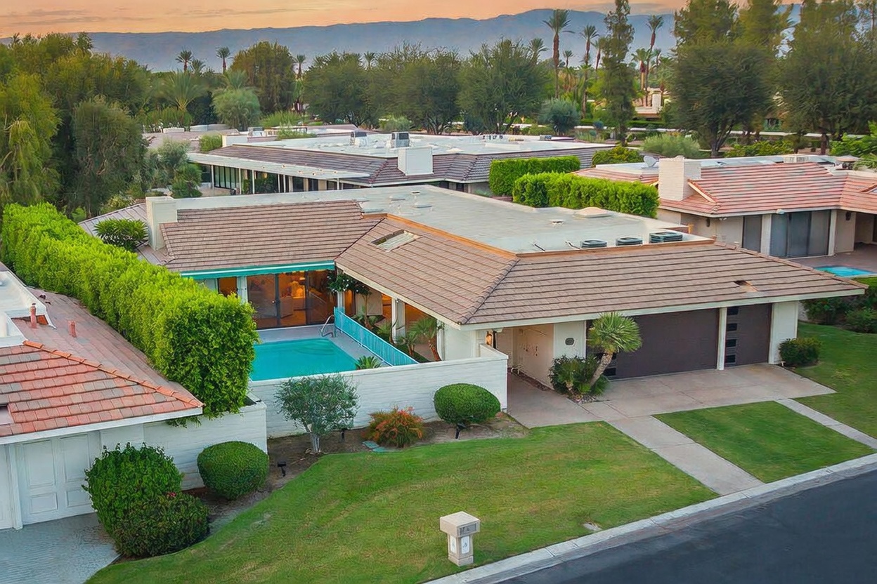 This elevated perspective captures a single-story residential property featuring a low-pitched tiled roof and a private swimming pool enclosed by a low wall. The home includes a multi-car garage and is surrounded by manicured lawns and mature landscaping, set against a backdrop of distant mountains at sunset. The architectural style reflects a classic desert-modern aesthetic common in the region.