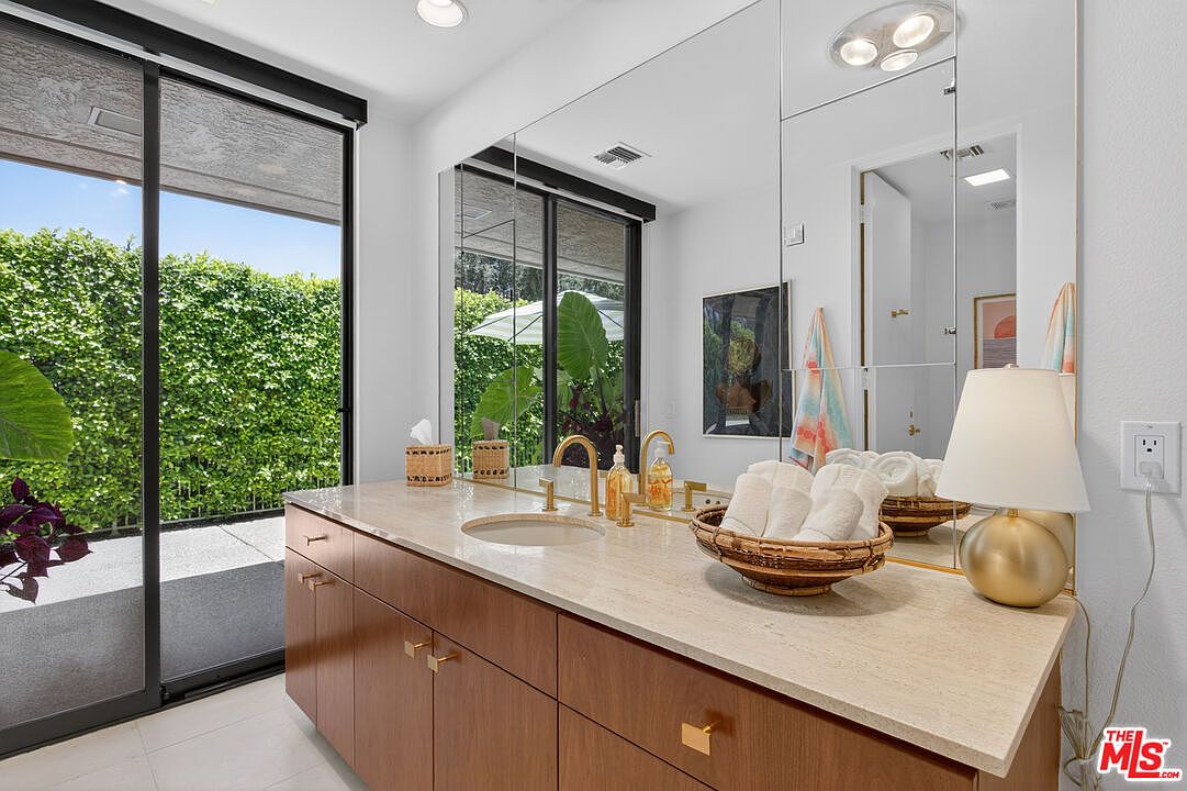 This bright primary bathroom features a warm-toned wood vanity topped with a light-colored stone counter and a gold-toned faucet. A large mirror spans the wall, reflecting the space and a sliding glass door that opens to a lush, green outdoor area. The room is accented with a gold table lamp and a basket of rolled white towels, creating a clean and inviting atmosphere.
