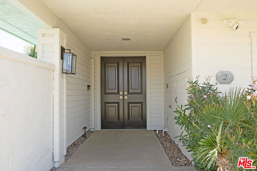 This entryway features a set of dark, double front doors set within a recessed, covered porch area. The exterior is finished with light-colored horizontal siding and a white brick wall on the left, complemented by a modern black wall-mounted lantern. A small, decorative plaque with the number '2' is mounted on the wall near a lush green shrub, creating a welcoming and private entrance.