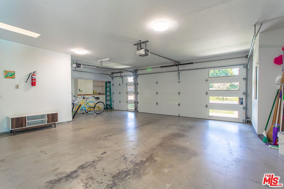 This spacious, well-lit two-car garage features a clean concrete floor and white walls, providing a bright and functional storage area. The space includes a modern white media console, a fire extinguisher mounted on the wall, and a pair of bicycles parked near a green ladder. The garage doors are equipped with windows that allow natural light to filter into the interior, creating an open and airy atmosphere.