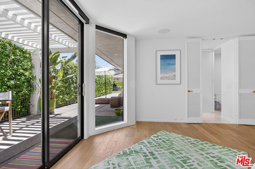 This bright bedroom features light wood flooring and a large sliding glass door that opens to a private patio area. A patterned green and white rug anchors the space, while a framed coastal print hangs on the white wall. The room connects to an adjacent bathroom through a set of white louvered doors, creating an airy and open atmosphere.
