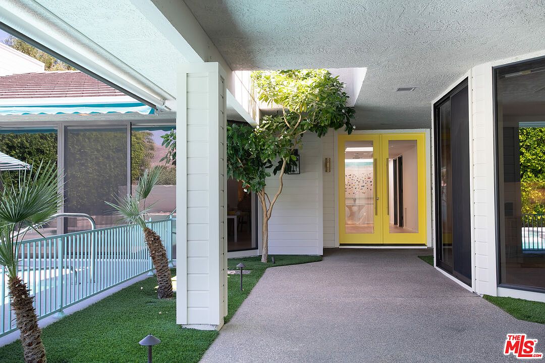 This bright and modern entryway features a striking pair of vibrant yellow double doors that serve as a focal point against the white exterior walls. A covered walkway with a textured concrete floor leads to the entrance, flanked by a small patch of artificial turf and a mature tree growing through a cutout in the roof. To the left, a light blue metal railing separates the walkway from a pool area, creating a clean, mid-century modern aesthetic.