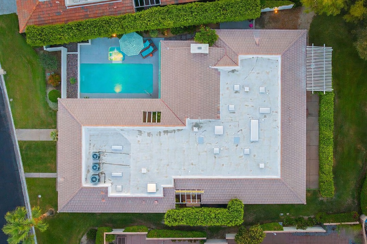 This top-down aerial view captures a residential property featuring a flat-roofed home with a tiled perimeter and a private swimming pool. The backyard includes a paved deck area with a patio umbrella and lounge chairs, surrounded by lush green lawns and manicured hedges. The perspective provides a clear layout of the roof, including multiple skylights and HVAC units, showcasing the home's footprint and outdoor living space.