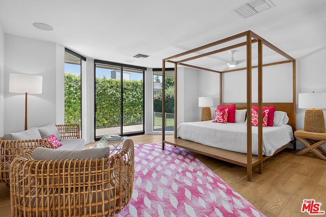 This spacious primary bedroom features a light wood canopy bed frame as the central focal point, complemented by a vibrant pink and white patterned area rug. The room is bright and airy, with large sliding glass doors that provide a view of the lush green hedge outside. A cozy seating area with woven chairs and a floor lamp adds a relaxed, inviting atmosphere to the space.