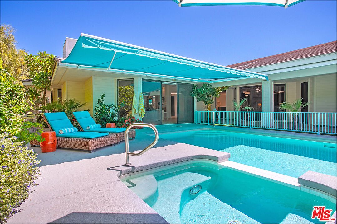 This bright, sun-drenched backyard features a private swimming pool and an integrated spa, both accented by a vibrant turquoise retractable awning. Two lounge chairs with matching cushions sit on the concrete deck, providing a relaxing space for outdoor leisure. The area is enclosed by a matching turquoise metal fence, creating a cohesive and inviting outdoor living space.