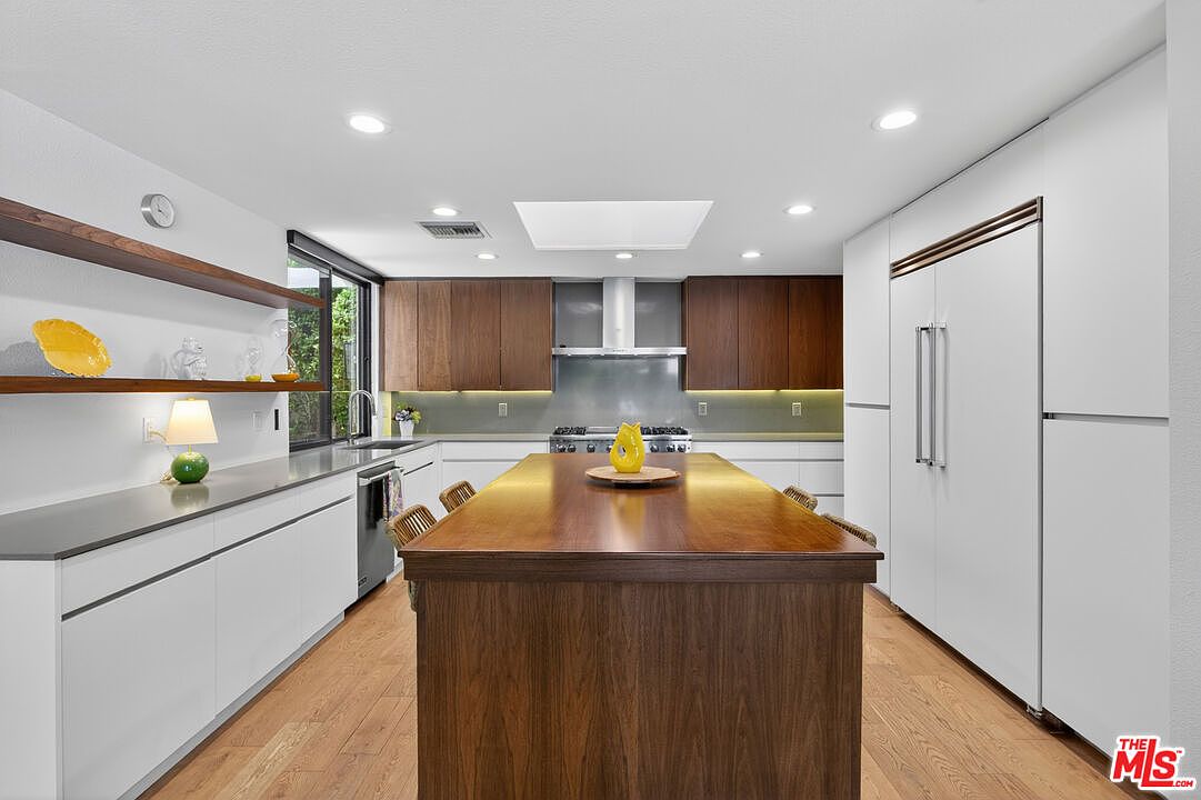 This modern kitchen features a large central island with a wood-toned base and a dark countertop, serving as the focal point of the room. The space is equipped with white cabinetry, stainless steel appliances, and open wood shelving, creating a clean and contemporary aesthetic. A skylight above the island provides natural illumination, complementing the recessed lighting throughout the bright, open-concept area.