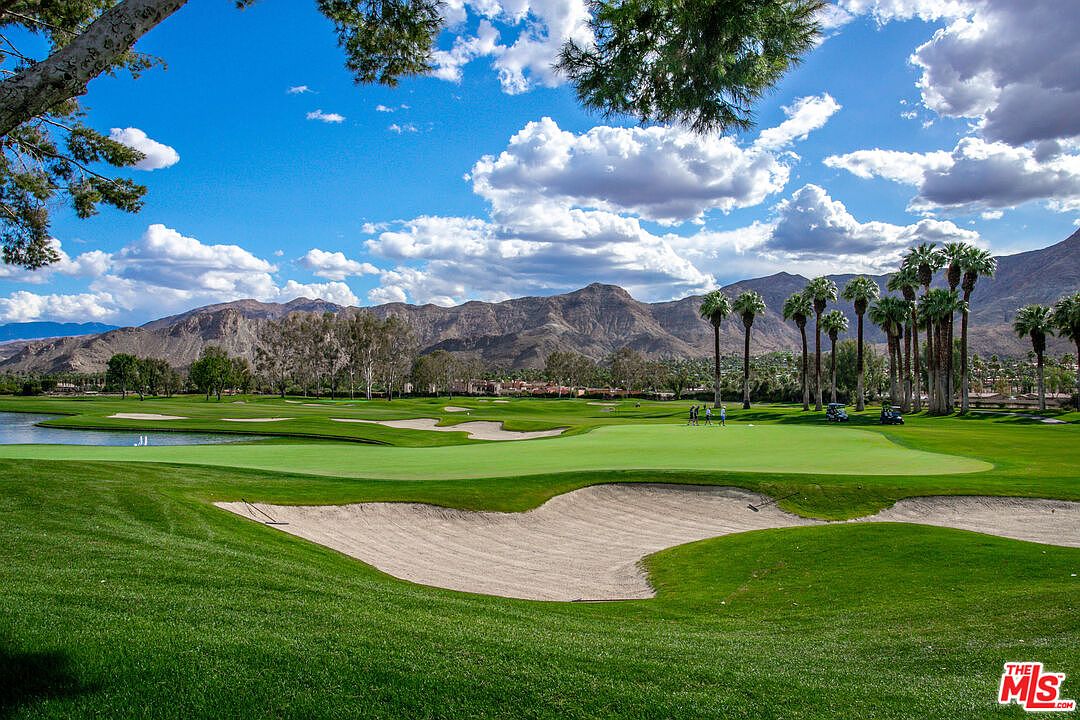 This image captures a scenic view of a manicured golf course, featuring a large putting green in the foreground with a sand bunker and a small pond to the left. In the background, a row of tall palm trees stands against a backdrop of rugged, sun-drenched mountains under a bright blue sky with scattered clouds. The perspective is a wide, eye-level shot that emphasizes the expansive, lush landscape and the tranquil atmosphere of the community.