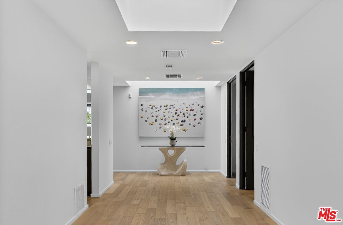 This bright, minimalist hallway features light wood flooring and crisp white walls, creating an airy and open atmosphere. A central console table with a unique sculptural base sits beneath a large, colorful piece of wall art, while a skylight above provides soft, natural illumination. The perspective is centered, drawing the eye down the corridor toward the artwork and the dark-framed doorways on the right.
