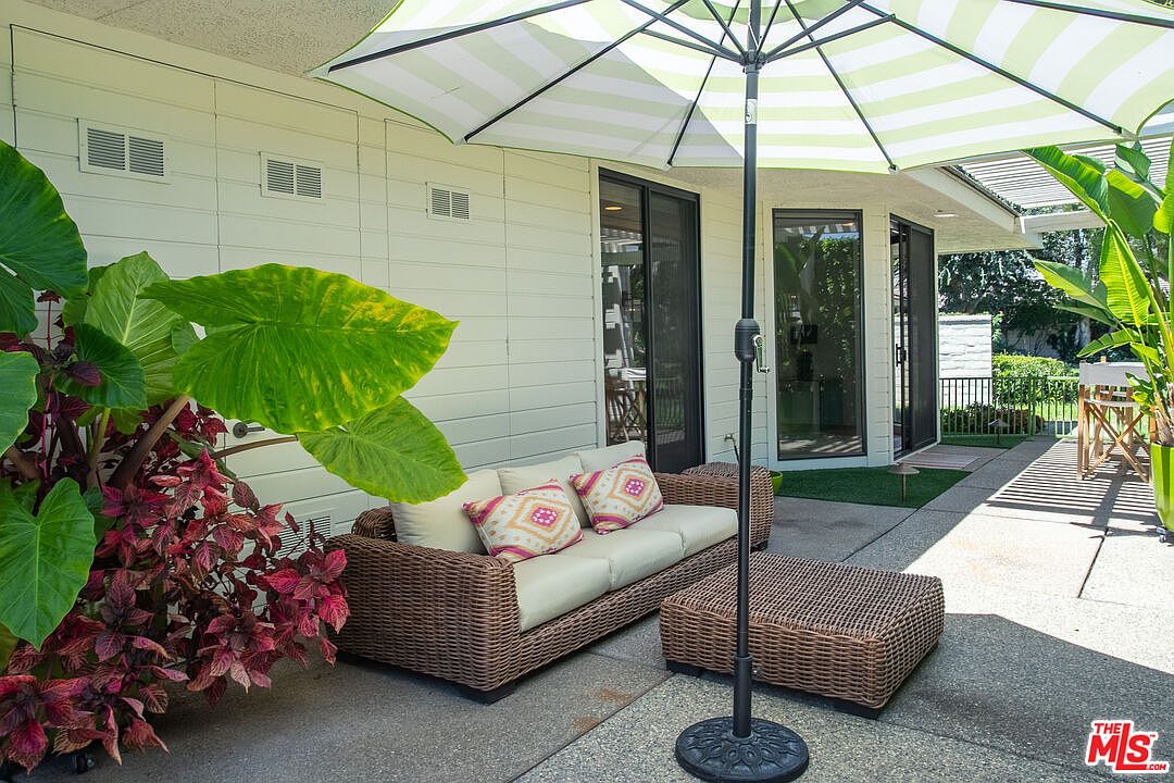 This inviting outdoor patio space features a comfortable wicker sofa with plush cushions and a matching ottoman, shaded by a large green and white striped umbrella. The area is set against a light-colored exterior wall with sliding glass doors that provide seamless indoor-outdoor access. Lush tropical foliage, including large-leafed plants and vibrant red-toned shrubs, adds a natural, serene touch to this relaxing backyard retreat.