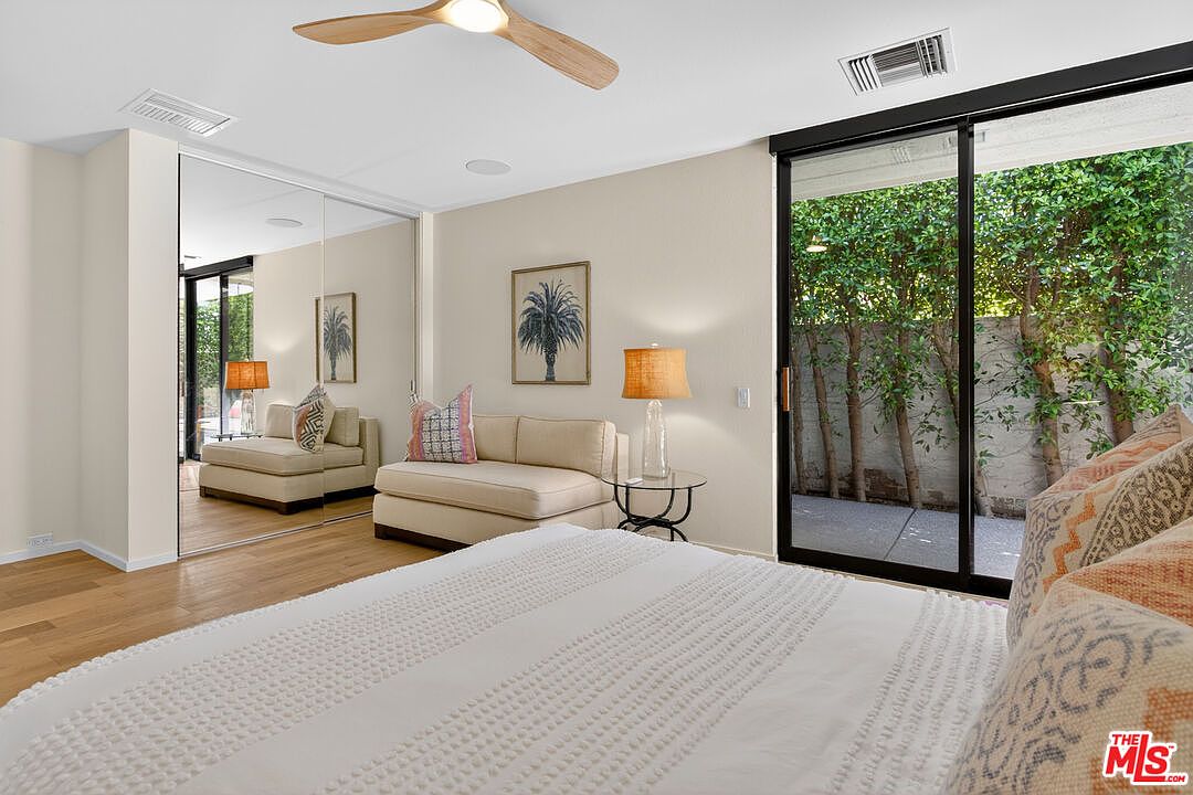 This bright and airy bedroom features light wood flooring and a neutral color palette, creating a serene atmosphere. A comfortable seating area with a sofa and side table is positioned against a wall of mirrored closet doors, while large sliding glass doors provide direct access to a private outdoor space. The room is illuminated by a modern ceiling fan and soft ambient lighting, offering a clean and inviting living space.