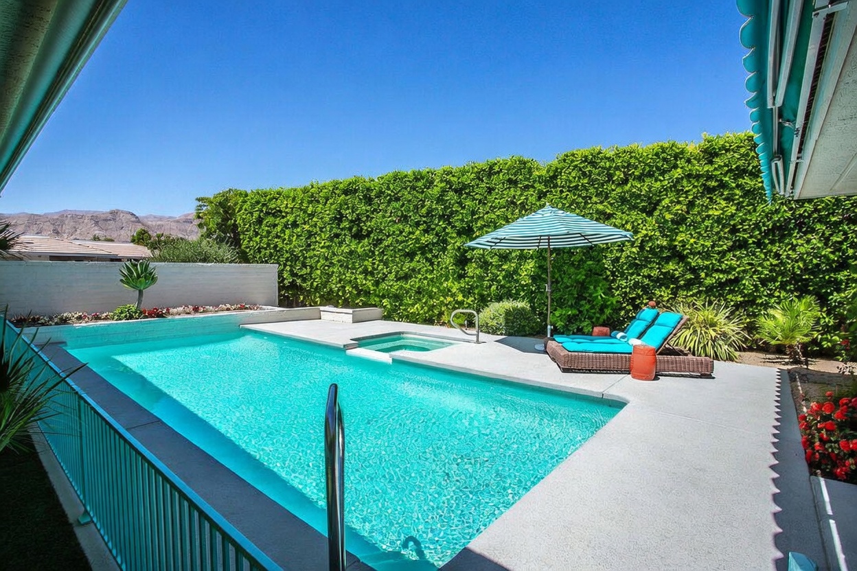 This bright, sun-drenched backyard features a rectangular swimming pool with an integrated spa, set against a backdrop of lush green hedges and distant mountains. A pair of turquoise lounge chairs sits under a striped umbrella on the concrete pool deck, creating a relaxing outdoor retreat. The perspective is captured from a covered patio area, looking out over the pool toward the privacy-focused landscaping.