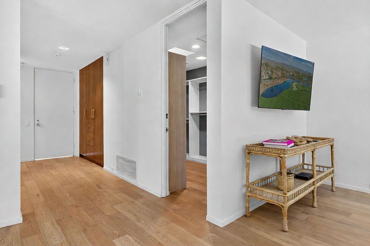 This interior view captures a bright, modern hallway featuring light wood flooring and crisp white walls. To the right, a rattan console table sits beneath a wall-mounted television, while a doorway leads into a walk-in closet with built-in shelving. A tall, dark wood cabinet provides contrast against the light-toned space, creating a clean and organized aesthetic.