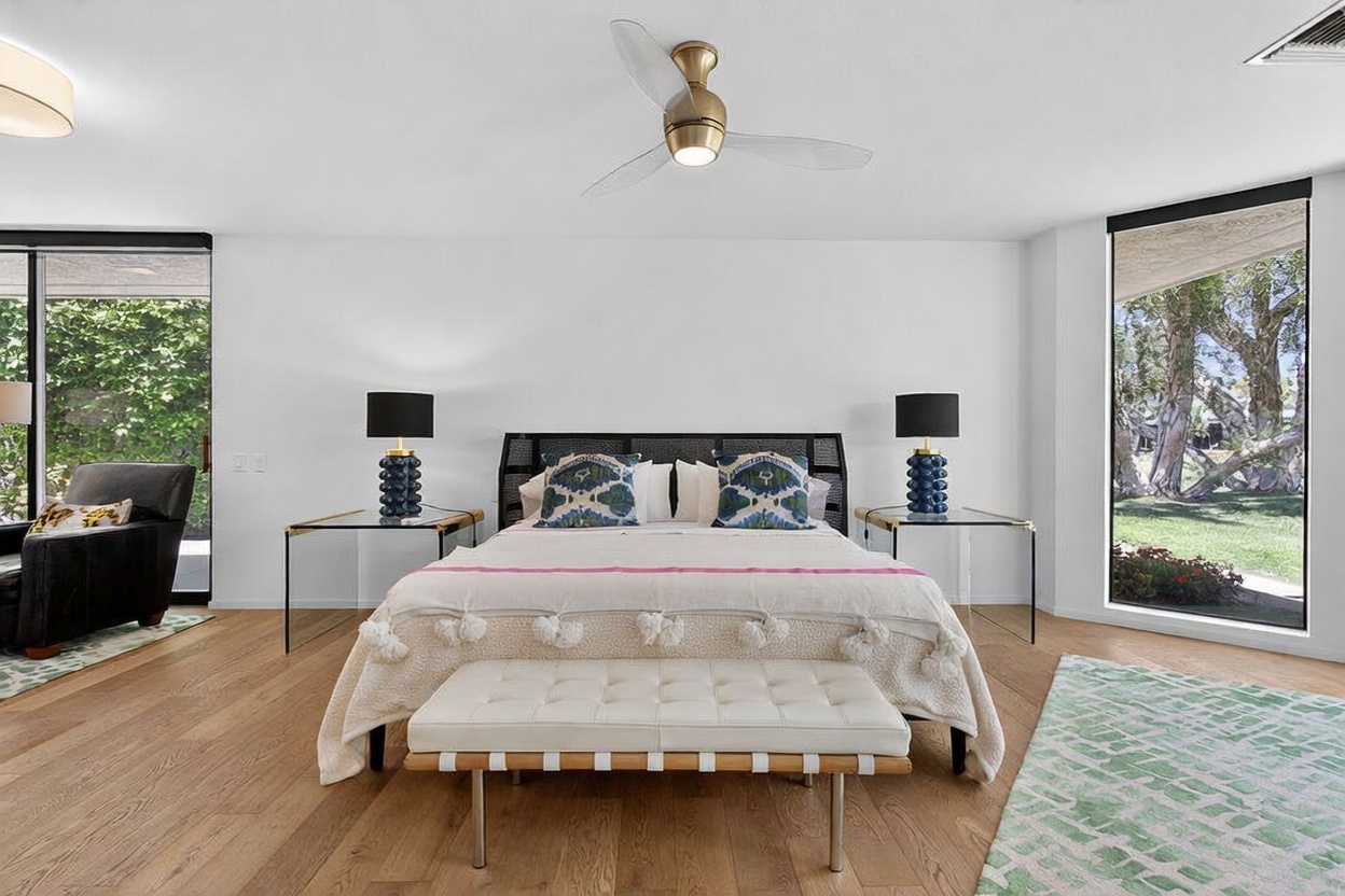 This spacious primary bedroom features a central bed with a dark, textured headboard, flanked by glass-topped nightstands and matching lamps. A white tufted bench sits at the foot of the bed, while large windows on either side provide abundant natural light and views of the surrounding greenery. The room is finished with light wood flooring, a modern ceiling fan, and a neutral color palette that creates a serene and airy atmosphere.