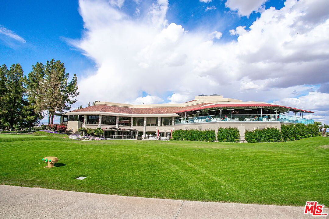 This wide-angle shot captures a large, multi-level clubhouse building situated on a meticulously manicured golf course. The structure features a prominent wrap-around terrace with glass railings, offering expansive views of the lush green lawn and surrounding landscape under a bright, cloudy sky. The perspective emphasizes the scale of the facility, highlighting its role as a central gathering point within the community.