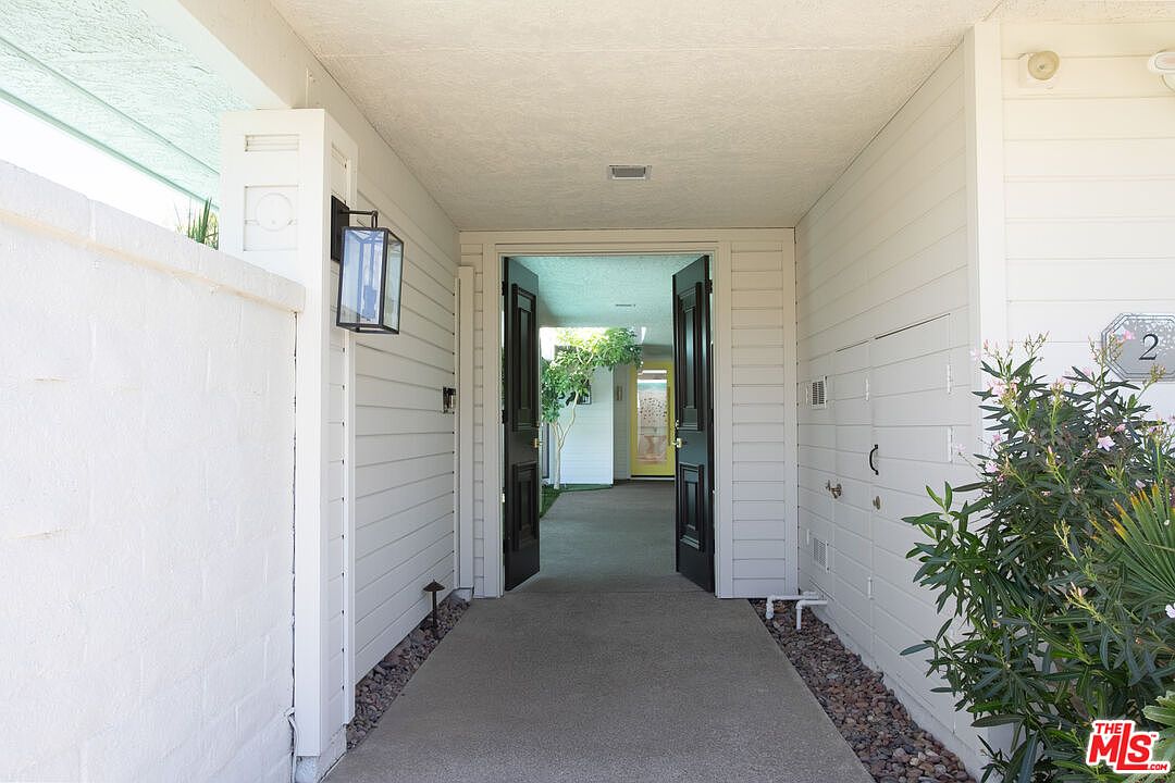This inviting entryway features a covered walkway leading to a set of open double doors, framed by white horizontal siding and a textured white wall. A modern black lantern-style light fixture illuminates the path, while a lush green plant adds a touch of nature to the right side of the entrance. The perspective looks through the open doorway into a bright, sunlit courtyard area, creating a welcoming and airy transition into the home.