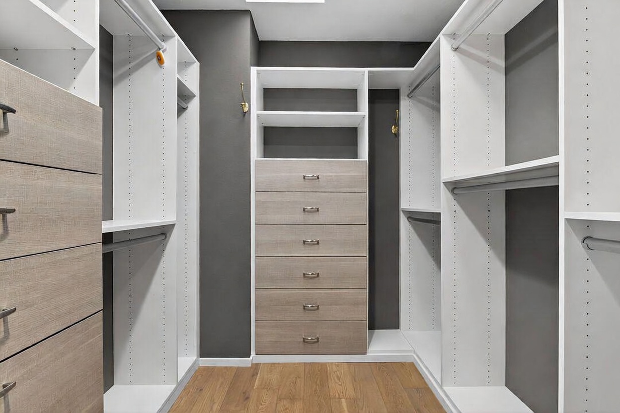 This organized walk-in closet features a symmetrical layout with light wood-toned drawers and white shelving units against a dark gray accent wall. The space includes multiple hanging rods, open shelving for storage, and a central bank of six drawers, all set atop warm-toned wood flooring. The perspective is centered, highlighting the clean, modern design and ample storage capacity of the custom closet system.
