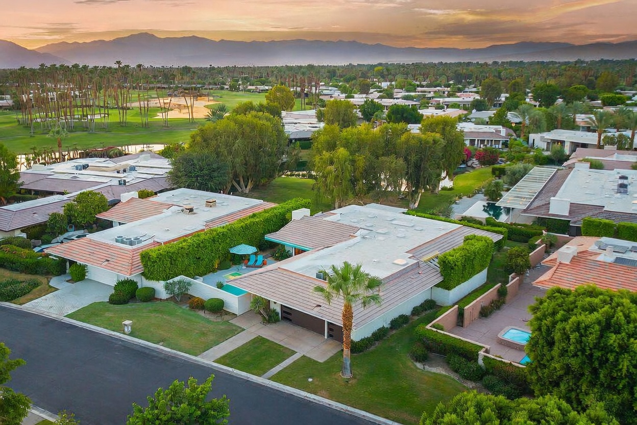This high-angle aerial view captures a residential property in a desert community, showcasing its flat-roofed architectural style and private courtyard. The home features a well-manicured lawn, a private pool, and lush landscaping, all set against a backdrop of a golf course and distant mountains at sunset. The perspective provides a comprehensive look at the neighborhood layout and the property's integration into the surrounding landscape.