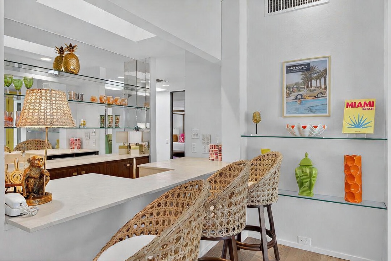 This interior view features a stylish wet bar area with a light-toned countertop and two woven bar stools. The space is accented by a mirrored backsplash, decorative pineapple sculptures, and a whimsical monkey-shaped lamp, while the adjacent wall displays framed art and colorful ceramic decor on glass shelving. The perspective captures a bright, well-lit transition space that leads toward a bedroom in the background, creating a playful and curated atmosphere.