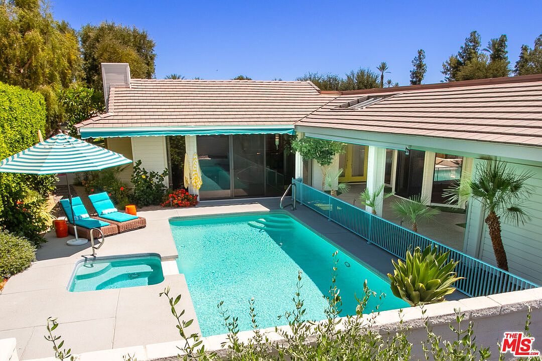This backyard scene features a rectangular swimming pool and an adjacent spa, set against a single-story home with a tiled roof. A turquoise safety fence surrounds the pool area, while a striped umbrella shades two lounge chairs on the concrete patio. The space is framed by lush greenery and a low wall, creating a private and inviting outdoor retreat.