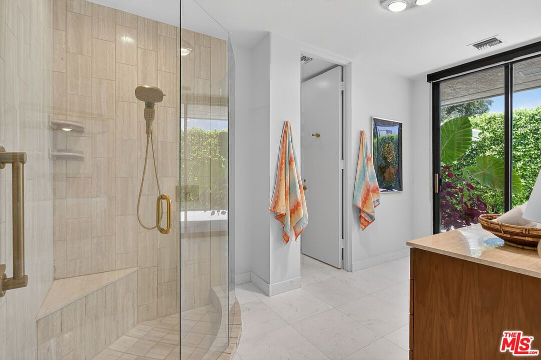 This bright bathroom features a walk-in shower with a glass enclosure, light-toned wall tiles, and a built-in bench. A wooden vanity with a stone countertop sits to the right, while large sliding glass doors provide natural light and a view of the lush greenery outside. The space is finished with light-colored floor tiles and two colorful towels hanging on the wall, creating a clean and airy atmosphere.