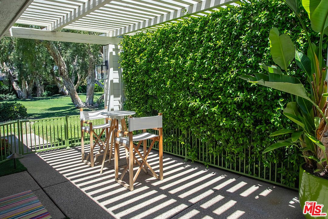 This inviting outdoor patio features a pergola overhead that casts dramatic, striped shadows across the concrete floor. The space is furnished with a small bistro table and two director-style chairs, positioned next to a lush, tall hedge that provides privacy. A green metal fence separates the patio from a grassy lawn and mature trees in the background, creating a serene and private retreat.