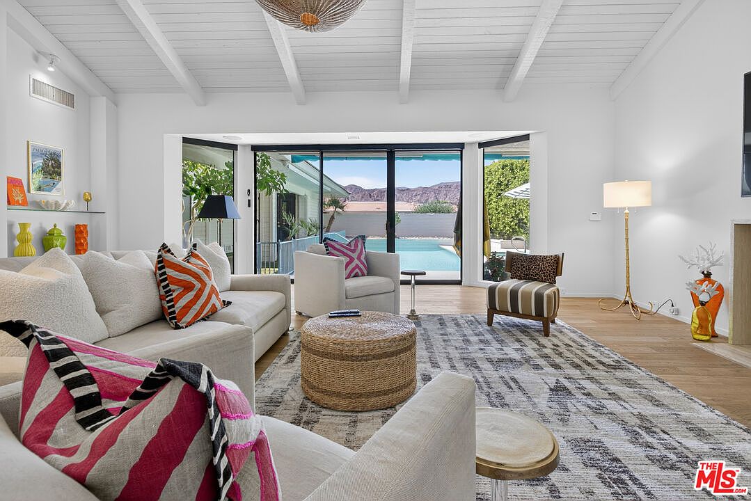This bright and airy living room features a vaulted white-beamed ceiling and a large sliding glass door that provides a clear view of the outdoor pool and mountain landscape. The space is furnished with a neutral-toned sofa accented by vibrant orange and pink pillows, a woven round ottoman, and a patterned area rug. A tall, gold-toned floor lamp and decorative accents add a touch of modern flair to this inviting, open-concept living area.