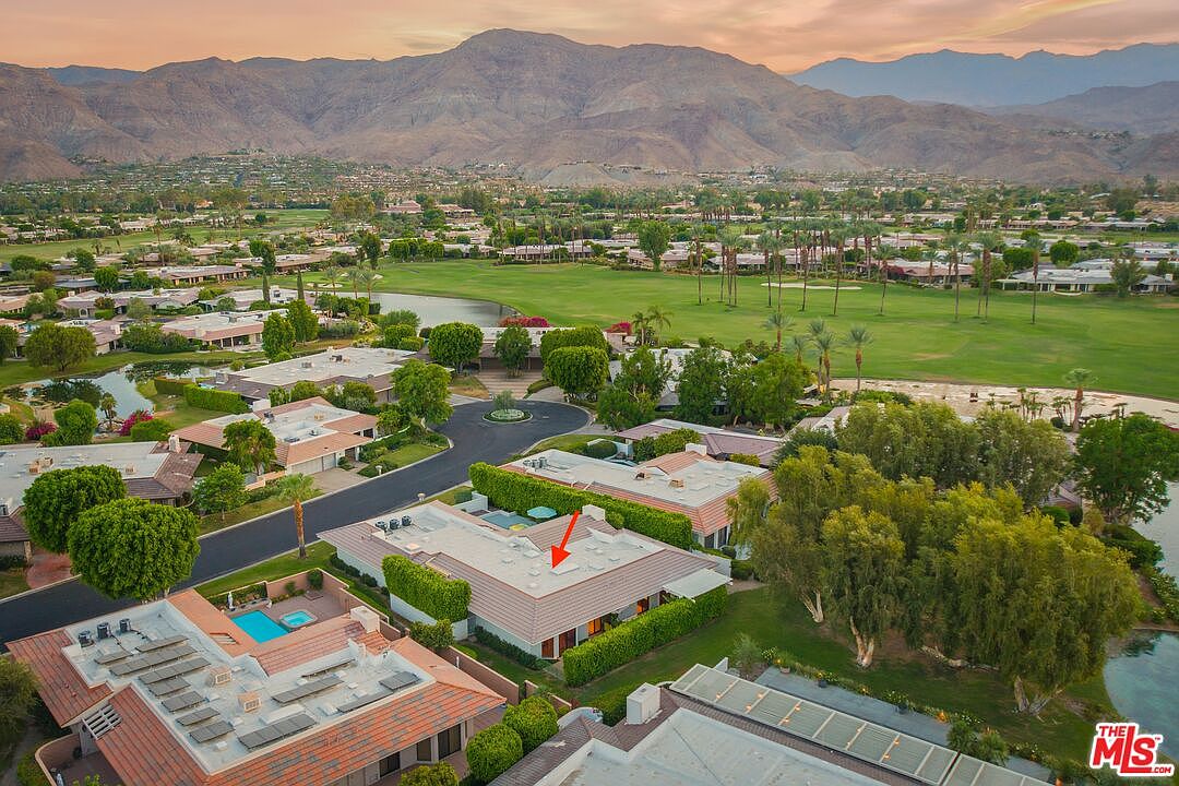 This high-angle aerial view captures a residential property nestled within a lush, manicured golf course community set against a backdrop of dramatic desert mountains. The home features a low-profile, multi-level roofline with a prominent red arrow indicating the specific residence, surrounded by mature trees, green fairways, and a nearby swimming pool. The perspective provides a comprehensive look at the neighborhood layout, emphasizing the serene and expansive outdoor environment.