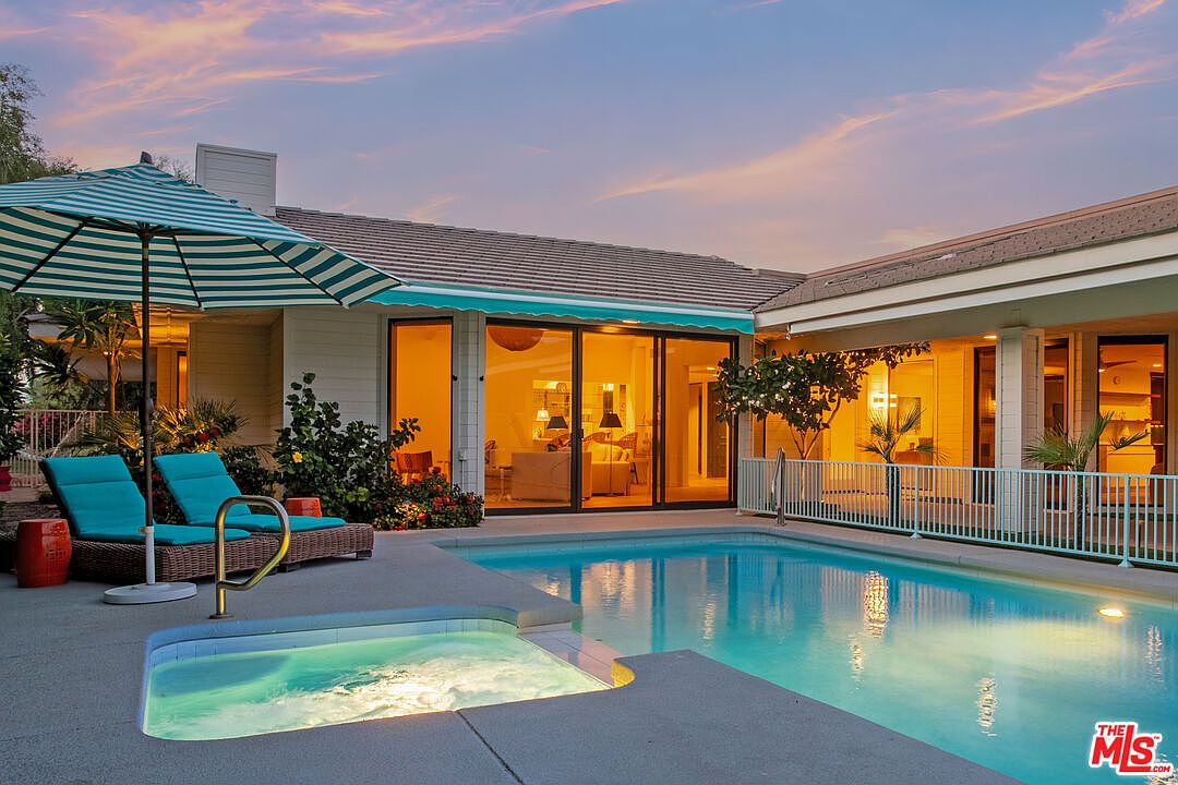 This backyard scene features a swimming pool and integrated spa set against a single-story home with large glass sliding doors. Two turquoise lounge chairs sit under a striped umbrella on the patio, while lush landscaping and a white metal fence frame the pool area. The warm, golden-hour lighting creates an inviting and serene atmosphere for outdoor relaxation.