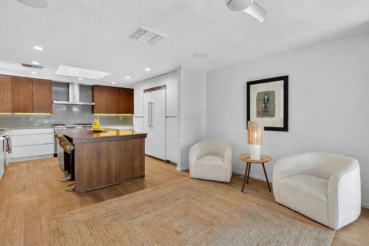 This open-concept space features a modern kitchen with dark wood cabinetry, a large central island, and stainless steel appliances. Adjacent to the kitchen is a cozy seating area with two cream-colored armchairs and a small side table, all set upon a large textured area rug. The room is bright and airy, characterized by light wood flooring and a neutral color palette that creates a welcoming, contemporary atmosphere.