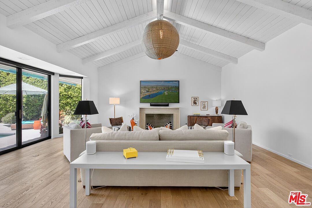 This bright and airy living room features a vaulted ceiling with exposed white beams and a large, textured pendant light fixture. A neutral-toned sofa is positioned in the center of the room, facing a fireplace with a stone surround and a wall-mounted television. Large sliding glass doors on the left provide a view of the outdoor pool area, while light wood flooring runs throughout the space.