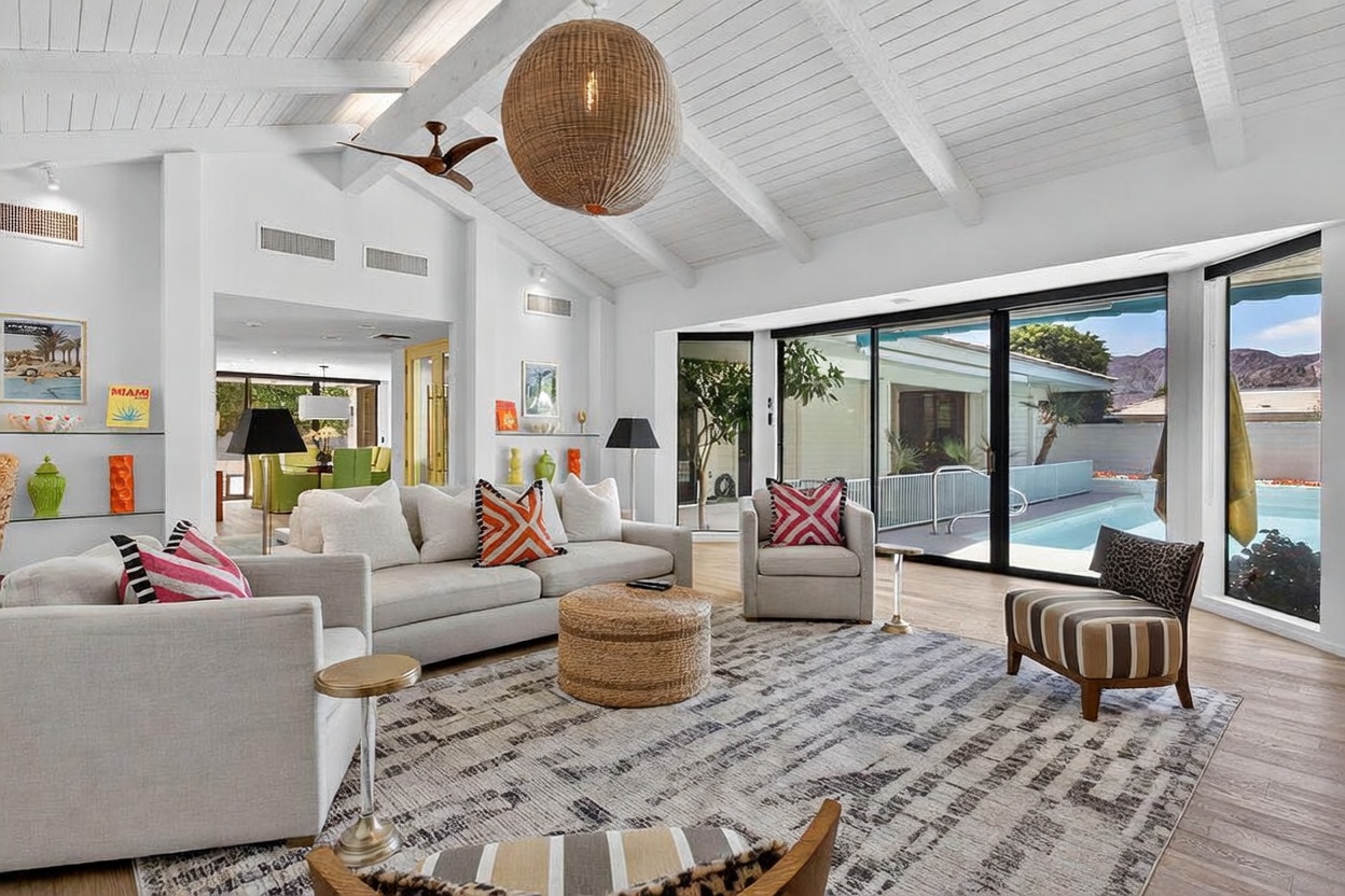 This bright and airy living room features a vaulted ceiling with exposed white beams and a large, textured pendant light fixture. The space is furnished with a neutral-toned sofa, patterned accent pillows, and a round woven ottoman, all set upon a large, textured area rug. Large sliding glass doors provide a seamless transition to the outdoor pool area, filling the room with natural light and offering a view of the surrounding landscape.