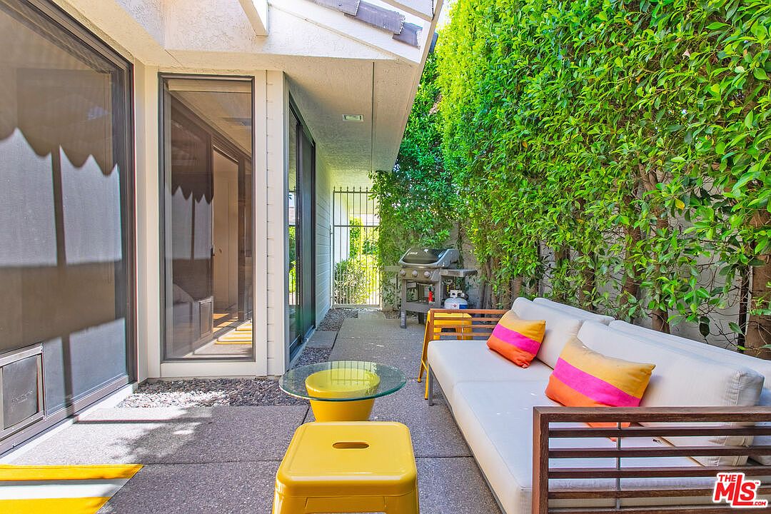 This inviting outdoor patio space features a modern sofa with vibrant striped cushions, complemented by yellow metal side tables and a glass-topped coffee table. The area is bordered by a tall, lush green hedge on one side and large sliding glass doors that provide seamless indoor-outdoor flow. A grill is positioned near the back gate, creating a functional and cozy setting for relaxation or casual dining.