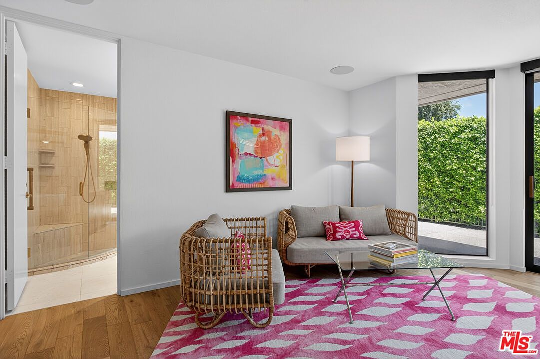 This bright living area features a contemporary seating arrangement with a rattan-style chair and a matching sofa, centered around a glass-topped coffee table on a vibrant pink and white patterned rug. A colorful abstract painting hangs on the wall, while a tall floor lamp provides warm lighting next to a large window overlooking lush greenery. The space flows directly into an adjacent bathroom, which is visible through an open doorway and showcases light-toned wall tiles and a glass-enclosed shower.