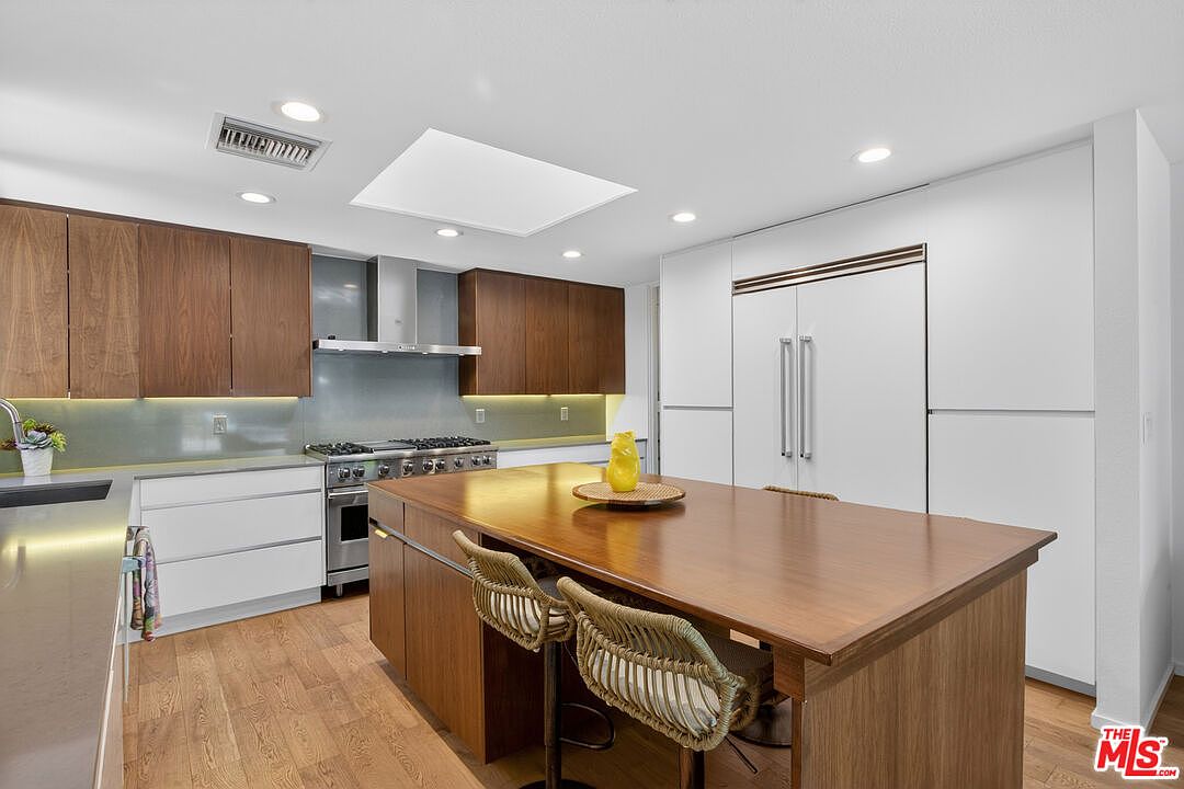 This modern kitchen features a large central island with a wood-toned surface and two woven bar stools, serving as a focal point. The space is equipped with stainless steel appliances, including a professional-style range, and is accented by a mix of dark wood and white cabinetry. Recessed lighting and a large skylight provide ample illumination, creating a bright and functional culinary environment.