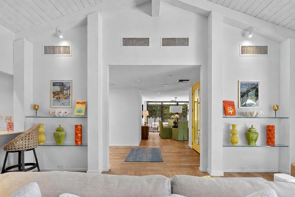 This bright and airy living space features a vaulted, white-painted wood-paneled ceiling and a central opening that leads to a dining area with a view of the outdoors. Symmetrical glass shelving units flank the central passage, decorated with colorful vases and framed artwork that add a vibrant, mid-century modern flair. The foreground includes a portion of a neutral-toned sofa, while the warm wood flooring creates a cohesive flow throughout the home.