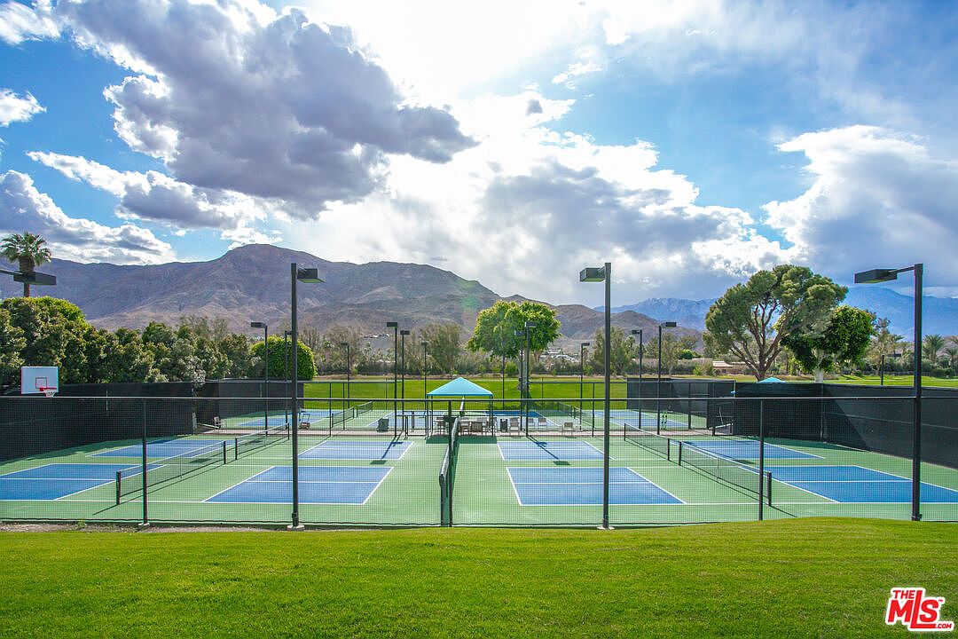 This image showcases a well-maintained community pickleball court facility set against a backdrop of dramatic mountains and a bright, cloudy sky. The scene features multiple blue-surfaced courts enclosed by black chain-link fencing, with a small shaded seating area in the center. The foreground consists of a lush green lawn, providing a clean, open perspective of the recreational space.
